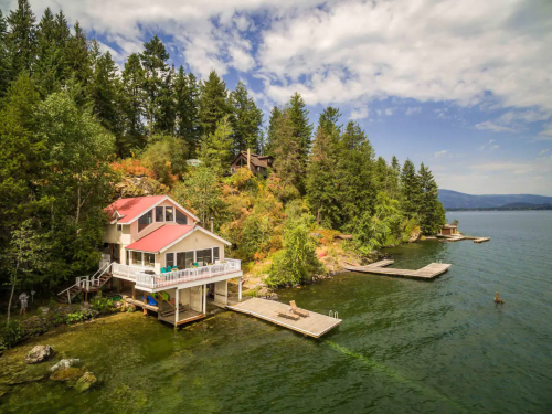 A scenic lakeside house with a red roof, surrounded by trees and docks extending into the water under a blue sky.